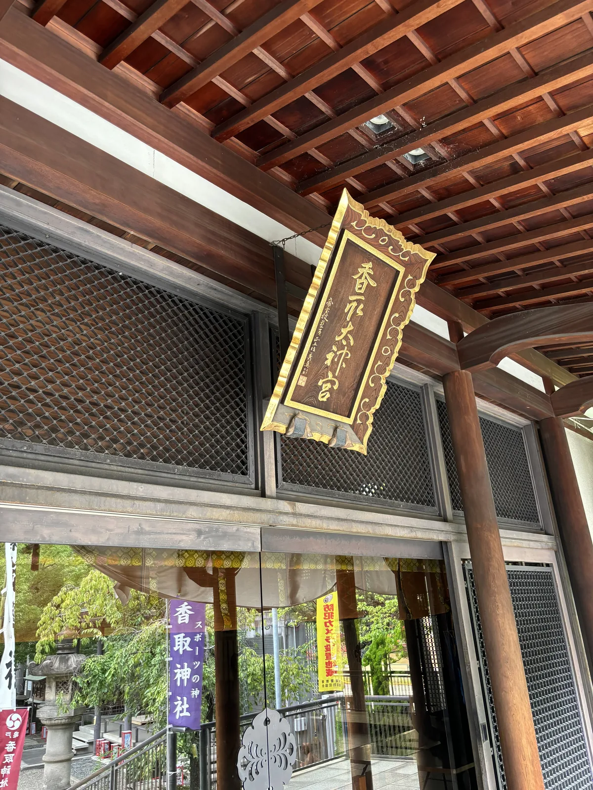從階下仰看拜殿簷下。一塊深色木底的扁額用鐵鍊吊著，外框是鎏金的唐草雕，額面金漆直書「香取太神宮」。額的左緣有一行小字「帝室技藝員　正五位　秀真」，下方一方朱印。背後是深紅色的椽與木格窗，再往下可以看見境內與寫著「スポーツ振興の神」的暖簾。