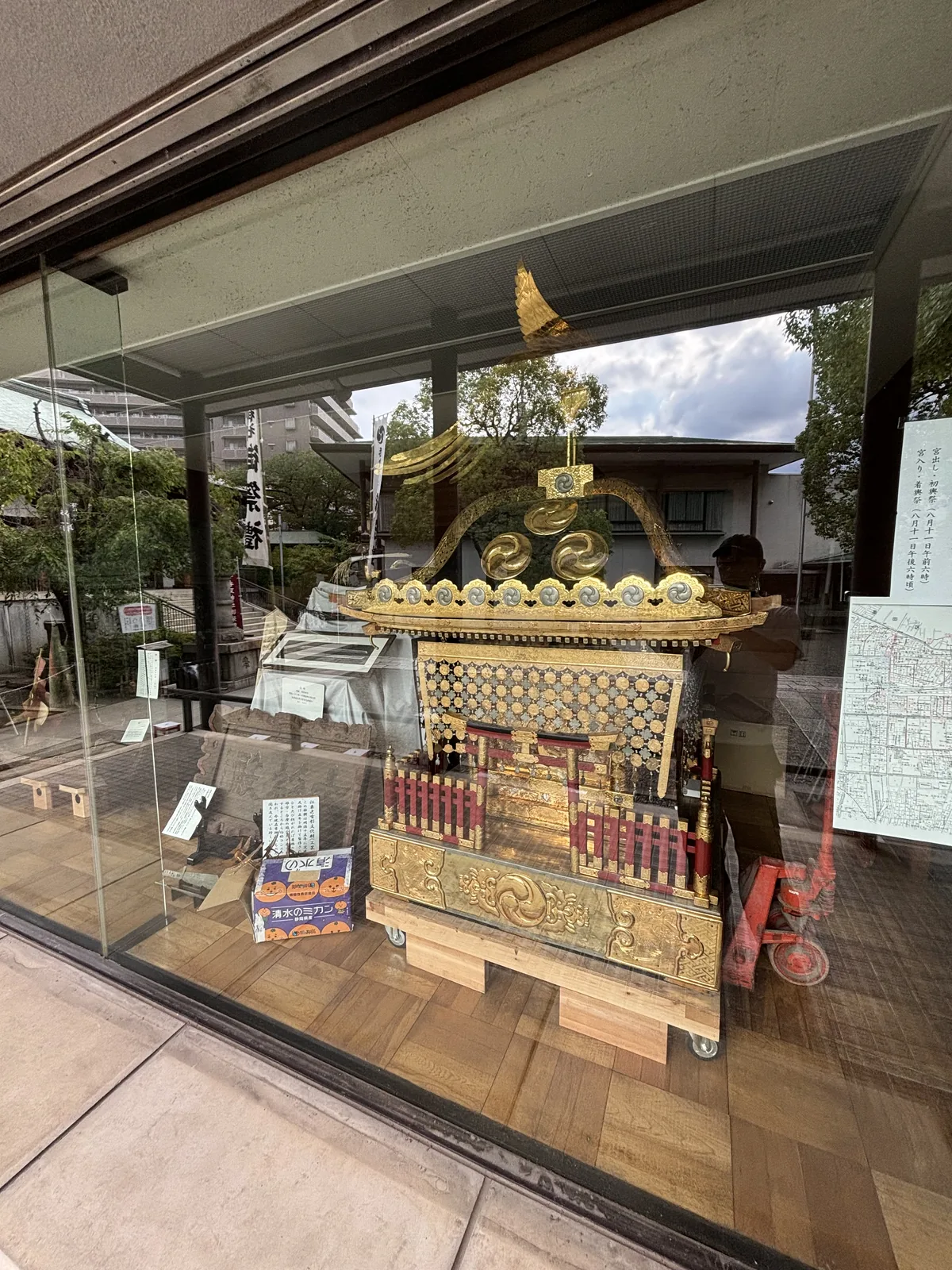 玻璃櫃裡的一頂大神輿。黑漆的輿身、金箔的裝飾與欄杆，屋頂中央立著一隻展翅的金鳳凰，底下架著木造的台座與轎槓，台座下墊著紅色的搬運台車。玻璃映出對面的境內建物與天空。