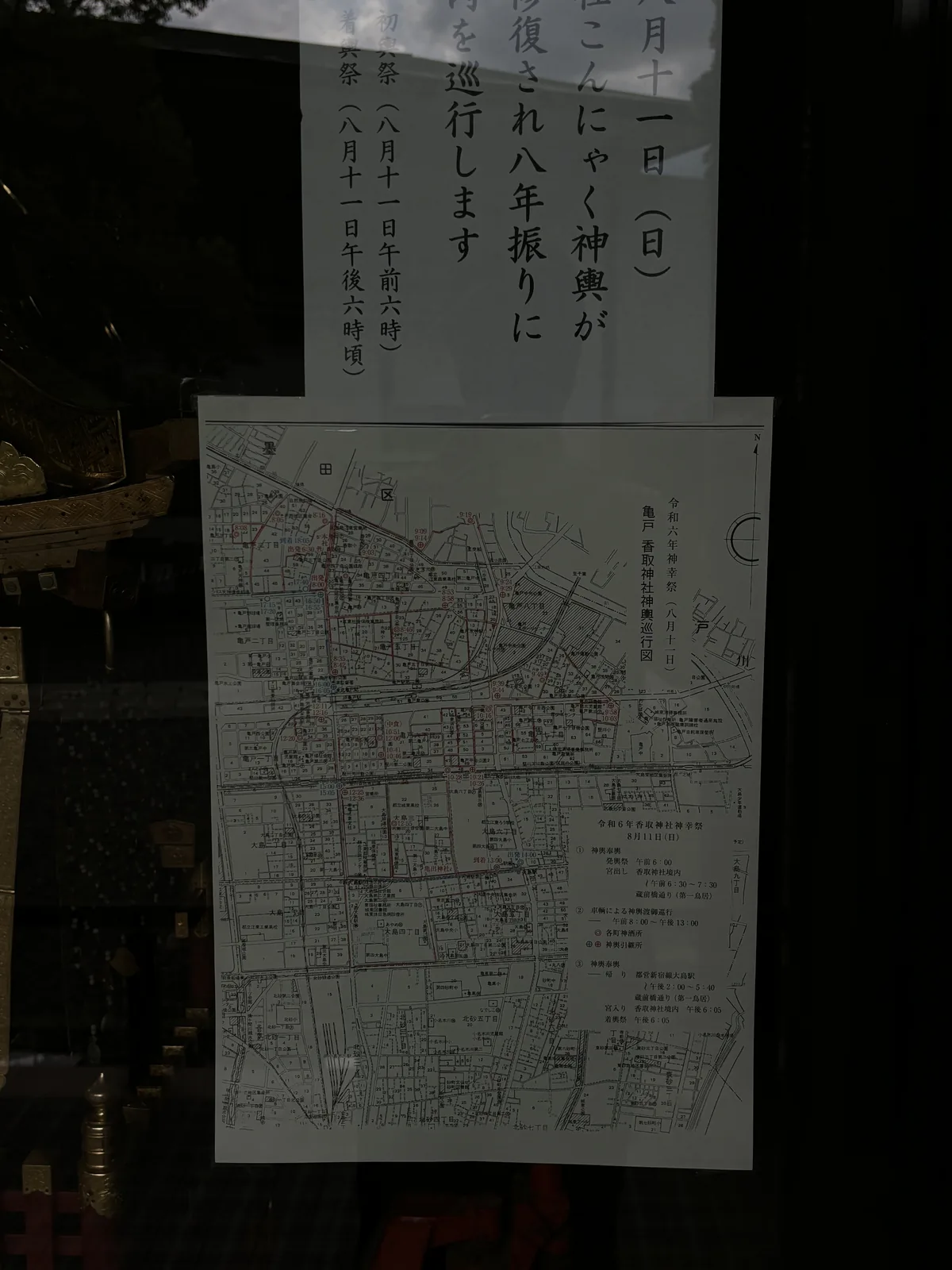 玻璃櫃的門上貼著兩張紙。上面一張是手寫的直書告示，可以讀到「八月十一日（日）」「こんにゃく神輿が」「復され八年振りに」「を巡行します」，以及「初輿祭（八月十一日午前六時）」「着輿祭（八月十一日午後六時頃）」。下面一張是印刷的巡行路線圖，街區上標著一連串時刻，圖的上方已經畫進墨田區。玻璃反射著樹影。