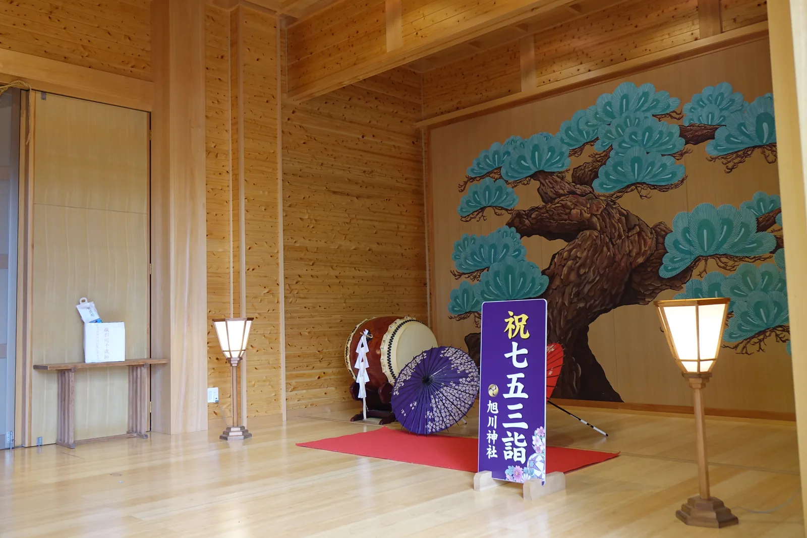 旭川神社神樂殿內部，全新的檜木壁面，正面畫著一株姿態橫張的松，地上鋪紅氈擺著太鼓與番傘