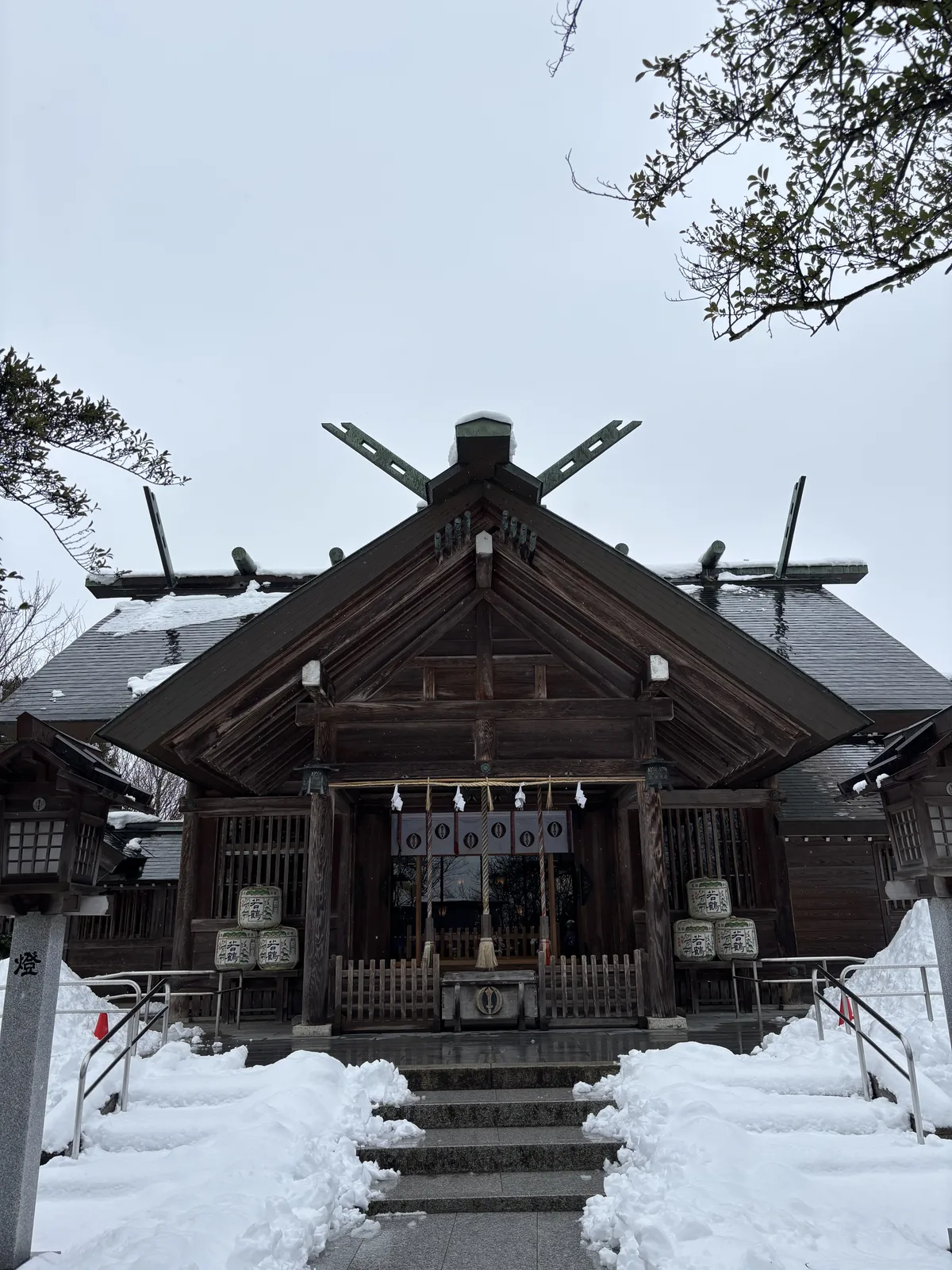 雪後的櫛田神社拜殿正面，素木切妻造，屋脊上交叉的千木與一排鰹木，向拜下垂著粗注連繩，兩側堆著奉納菰樽