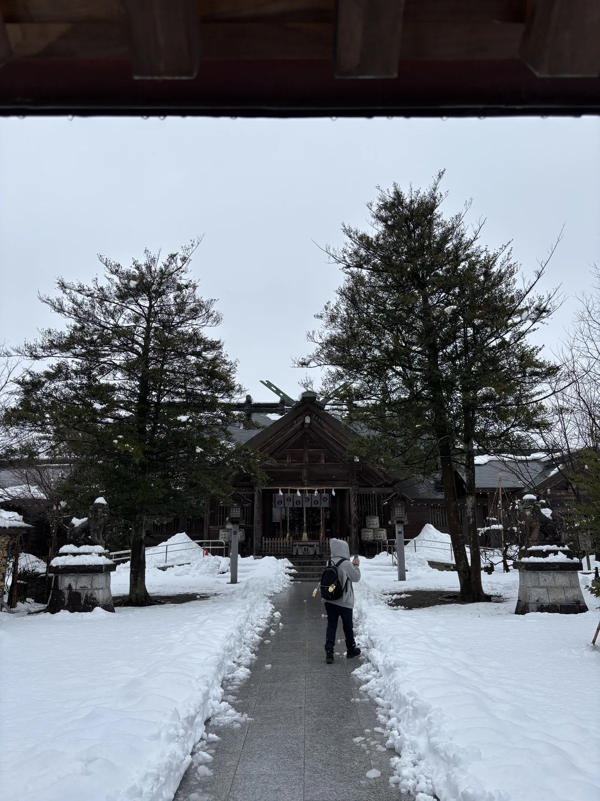 從境內一處簷下望向櫛田神社的拜殿，石板路兩側是剷開的雪，左右各一座高石座上的狛犬與木造燈籠