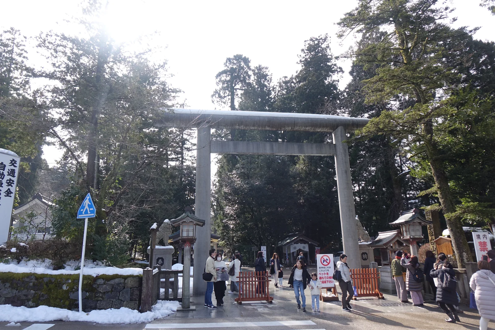 白山比咩神社境內入口的石造明神鳥居，笠木上積著一層雪，柱腳有斑馬線橫過，左側白幟寫著交通安全