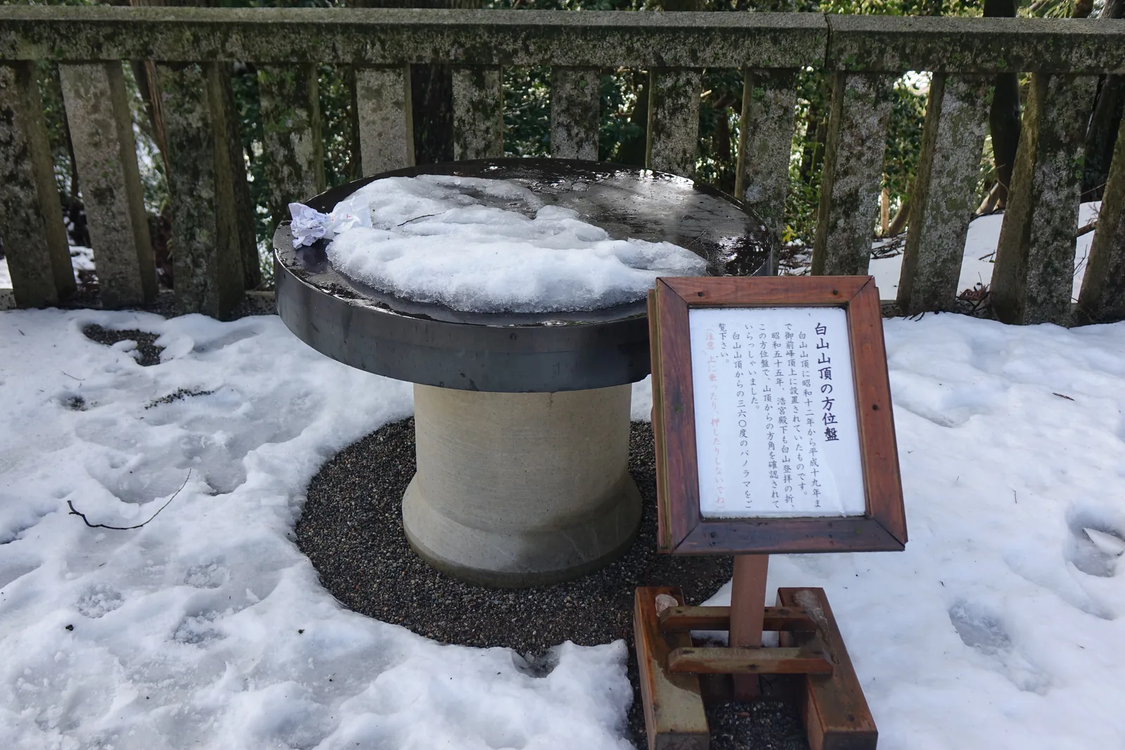 遙拜所的方位盤，圓形金屬盤面蓋著雪與冰，旁邊木框裡的手寫立札說明它原設在白山御前峰頂上