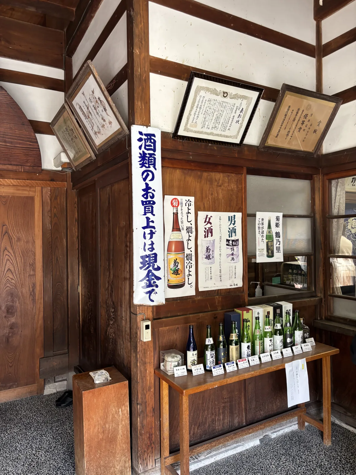 菊姫的店內，白牆與木柱之間掛滿獎狀與表彰狀，長桌上排著酒瓶與手寫價牌