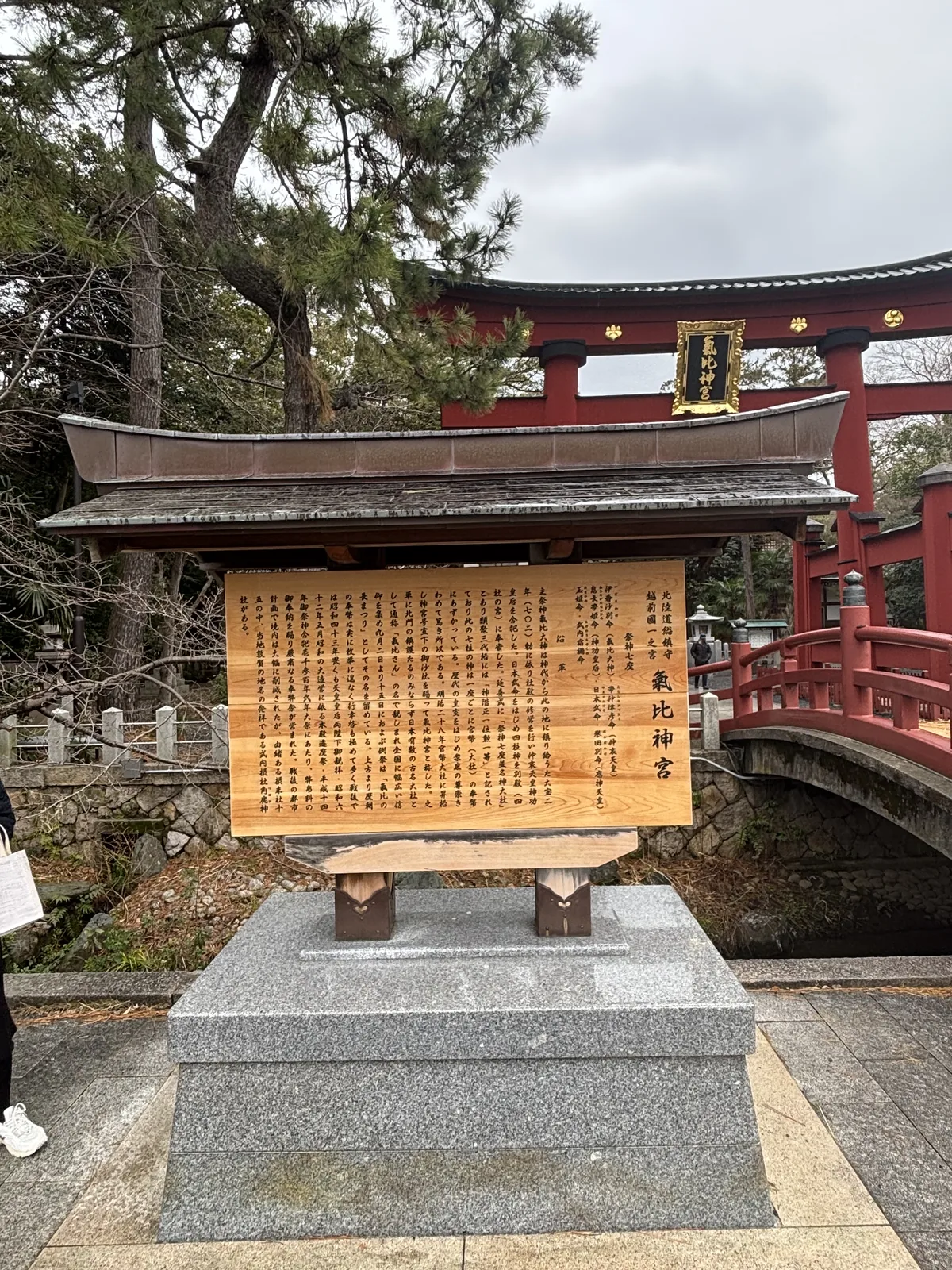 氣比神宮的由緒板，木造覆屋下的欅木一枚板，背後是大鳥居與朱塗神橋