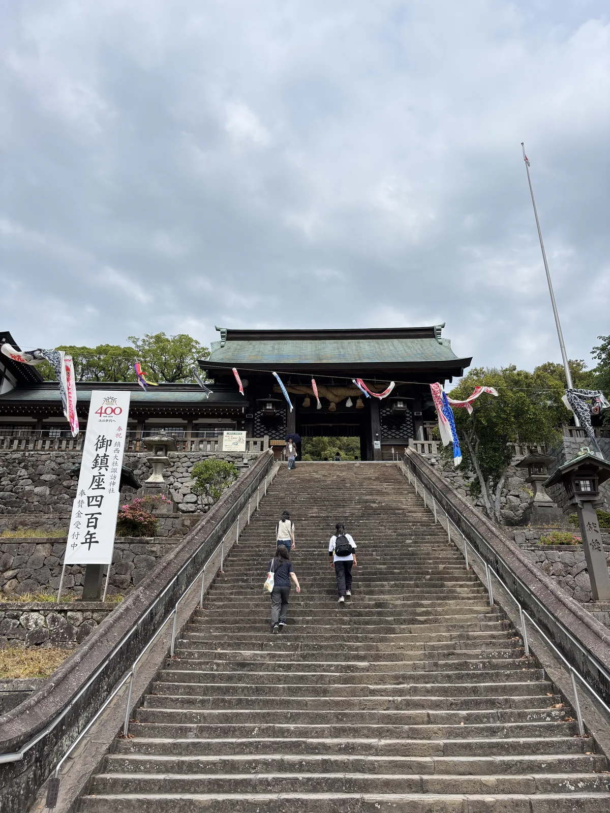 長崎諏訪神社的長石階，盡頭是黑色木造、青綠色屋頂的大門，石階左側立著寫有「御鎮座四百年」的白幟
