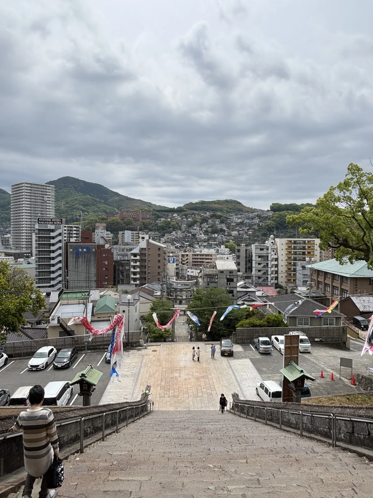 從諏訪神社的石階頂端往下看長崎市街，山坡上密集的房屋一直延伸到遠處的山，參道上空橫掛著鯉幟