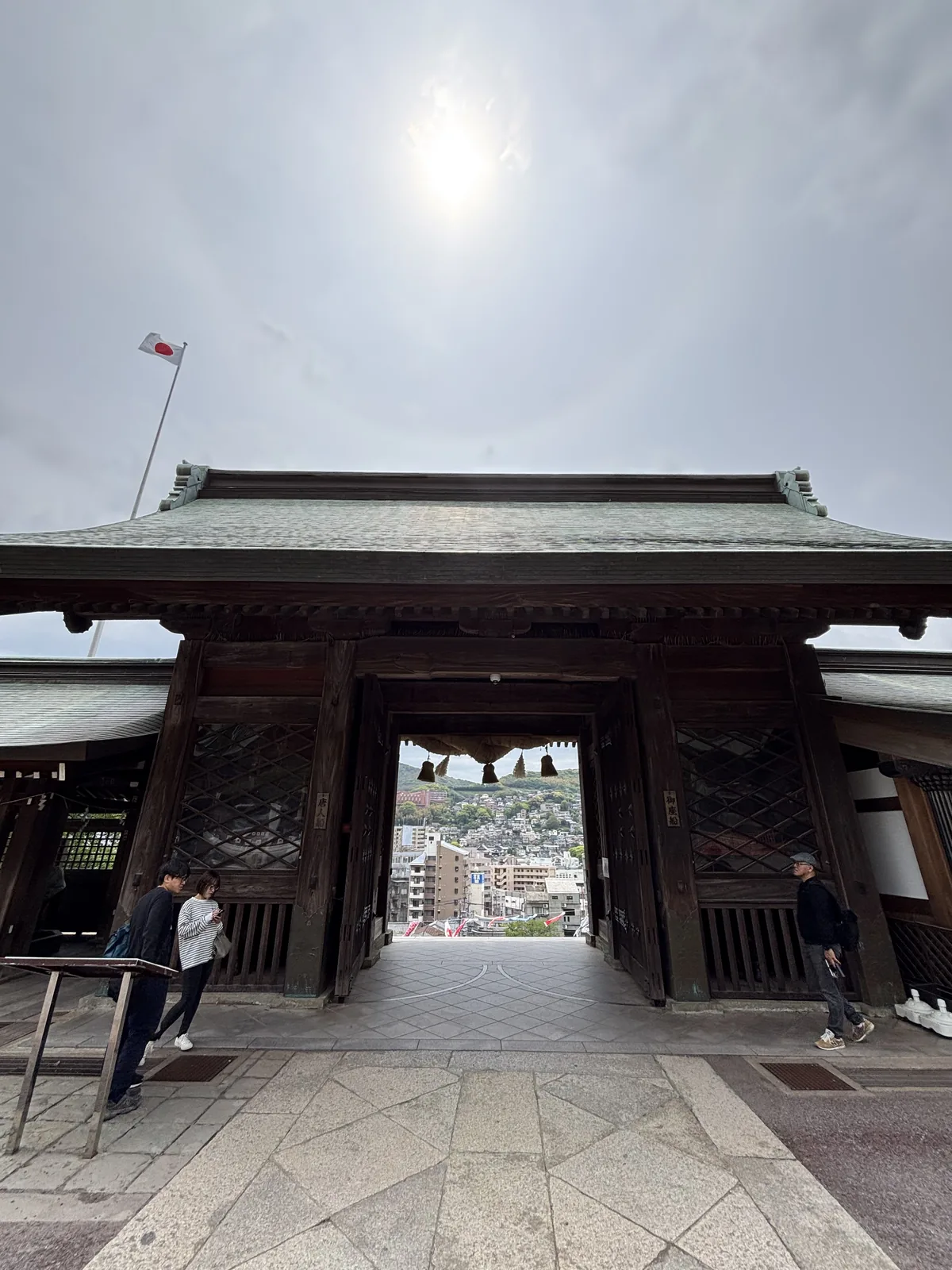 從諏訪神社大門內側往外看，門洞正好框住長崎市街與遠山，門楣下垂著三束注連繩，太陽在門的正上方