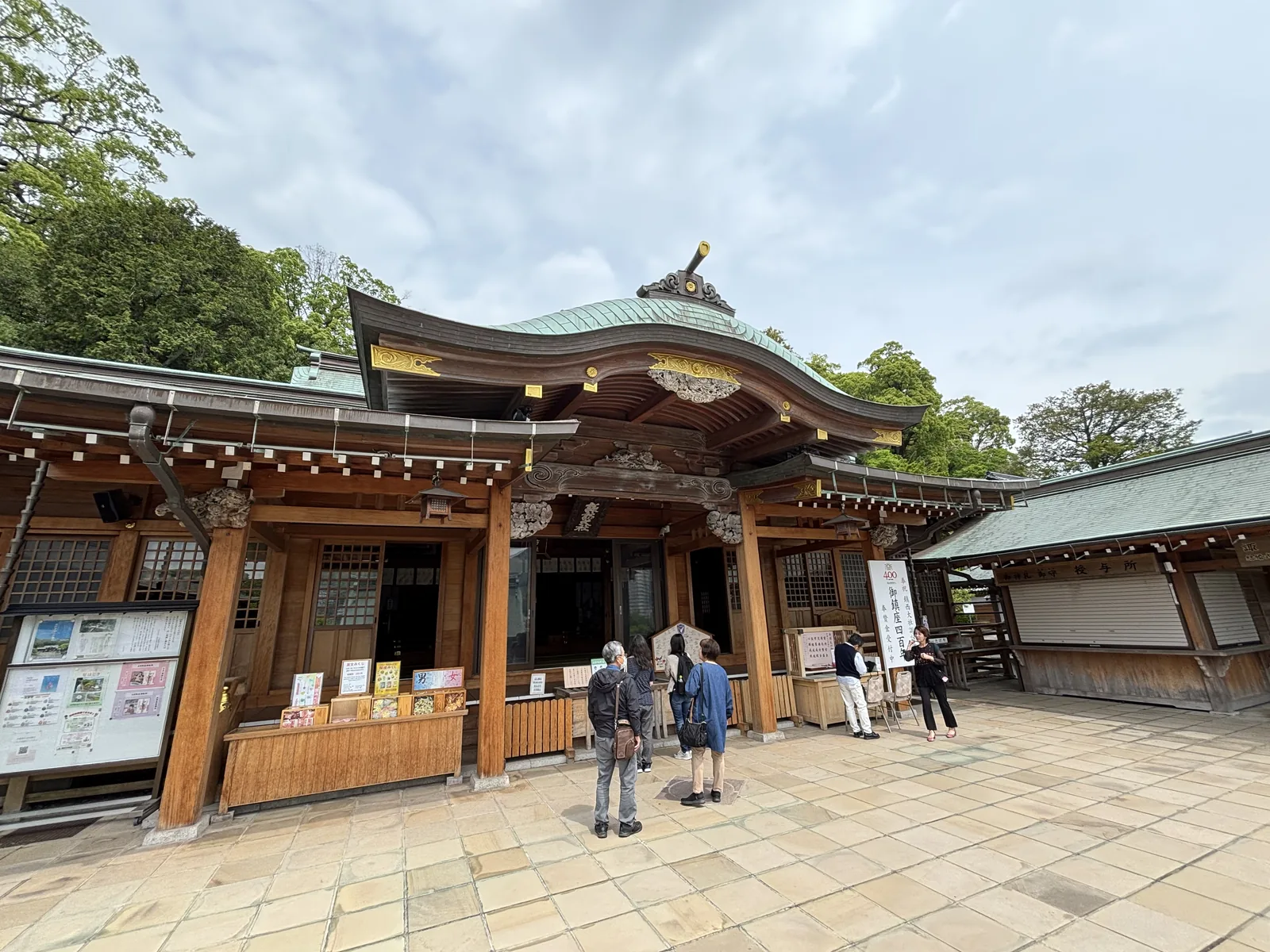 諏訪神社的授與所，木造平屋、青銅色屋頂帶唐破風，正面陳列御守與繪馬，幾位參拜者站在前面