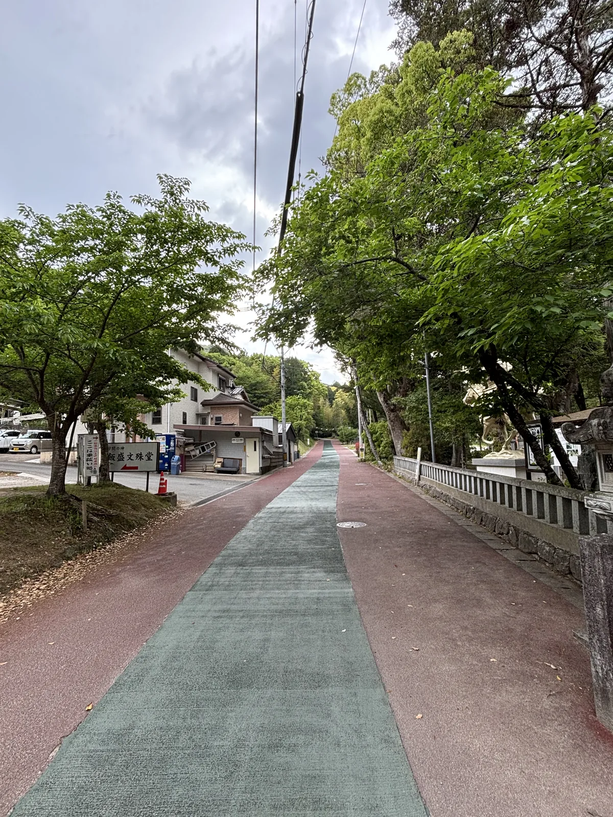 飯盛神社門前的道路，綠色與紅色分色鋪面的人行道往前延伸，右側是神社的石欄杆與樹木，左側是住宅