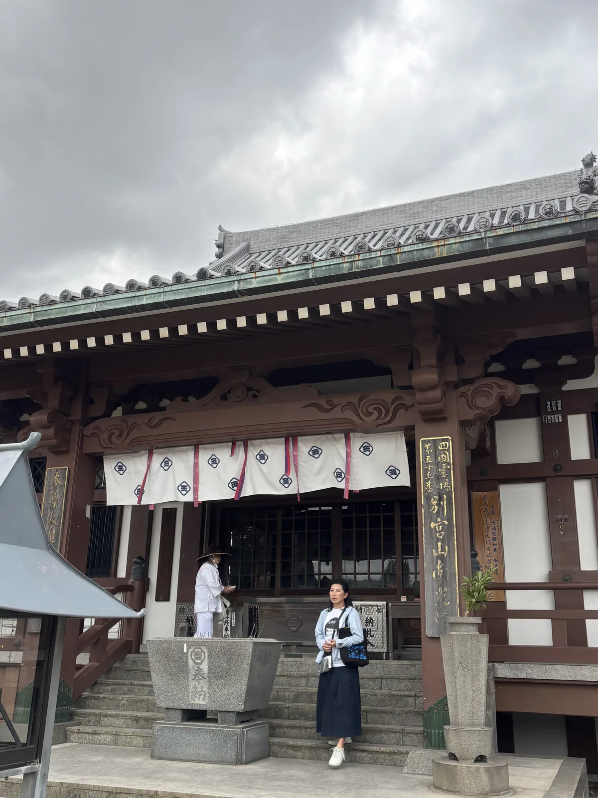 南光坊本堂正面：朱褐色的木構，向拜（本殿正面屋簷向外伸出、供參拜者站立的抱廈）下垂著一排白幕，幕上印著角切折敷裡三道橫畫的紋，柱上掛著黑底金字的木札，石階上站著一位戴菅笠、穿白衣的遍路