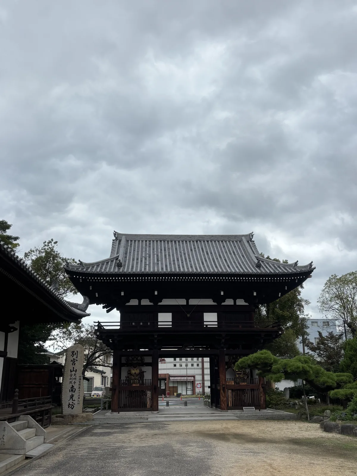 南光坊山門，從境內往外拍：二重門的左右門洞裡各站一尊披甲、背後有光背的天部像，門外是國道與對街的商業建築，門左側立著刻「別宮山　光明寺　金剛院　南光坊」的石標