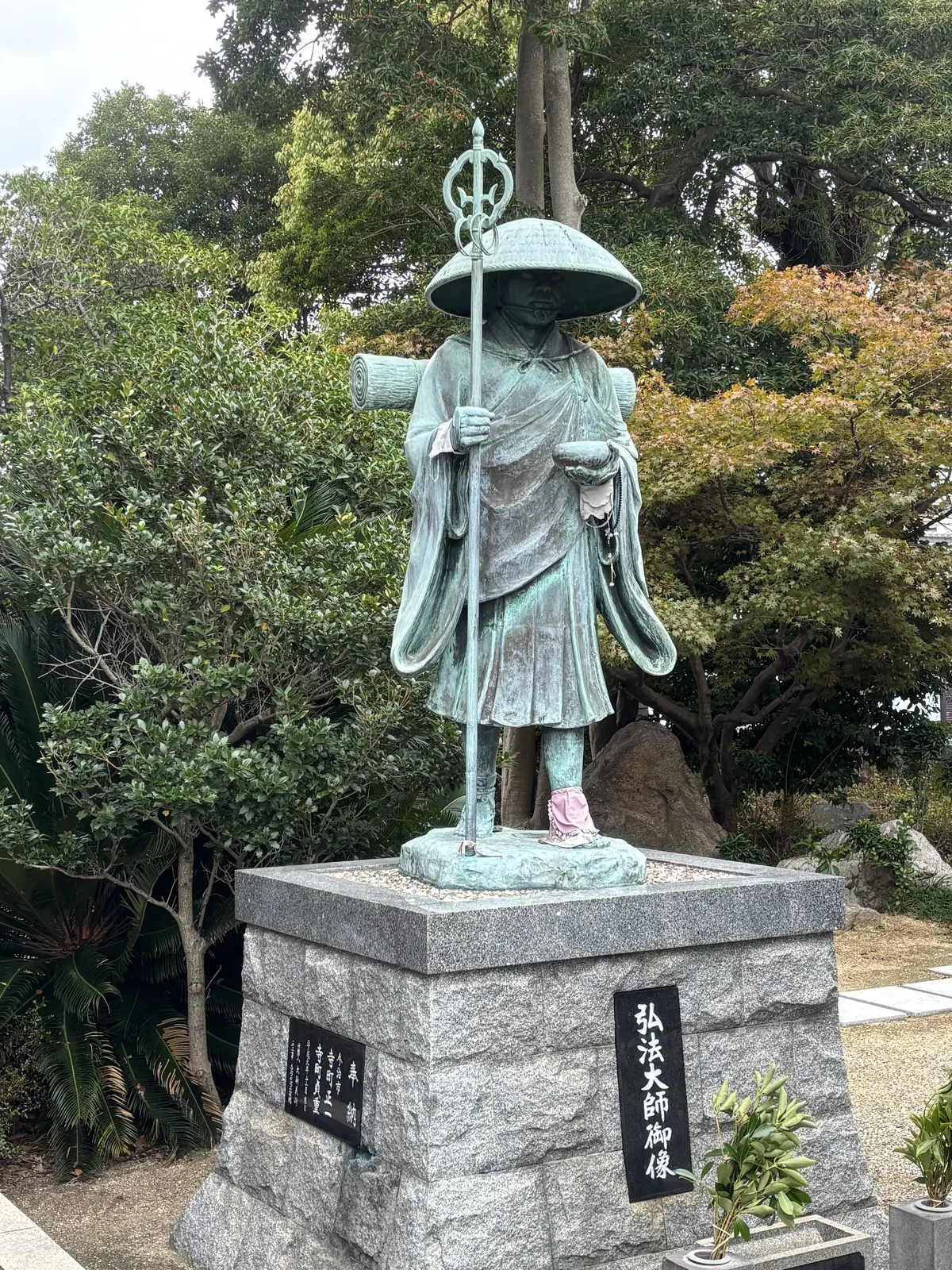 南光坊境內的弘法大師御像：青銅立像戴笠、持錫杖、托鉢，立在御影石的基壇上，基壇正面刻「弘法大師御像」，像的腳上繫著一條粉紅色的布