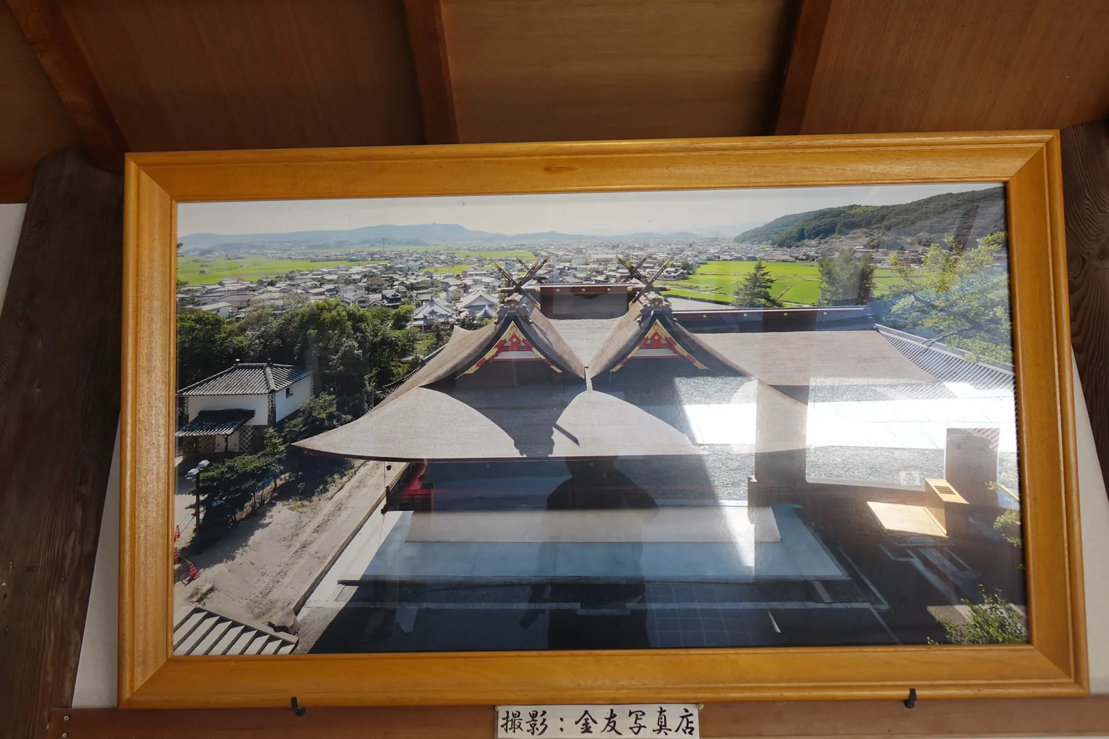 境內展示的空拍照片，框在木框裡：從正上方看，本殿兩個破風之間的棟接成片假名「エ」的形狀，後方是吉備津的聚落與水田