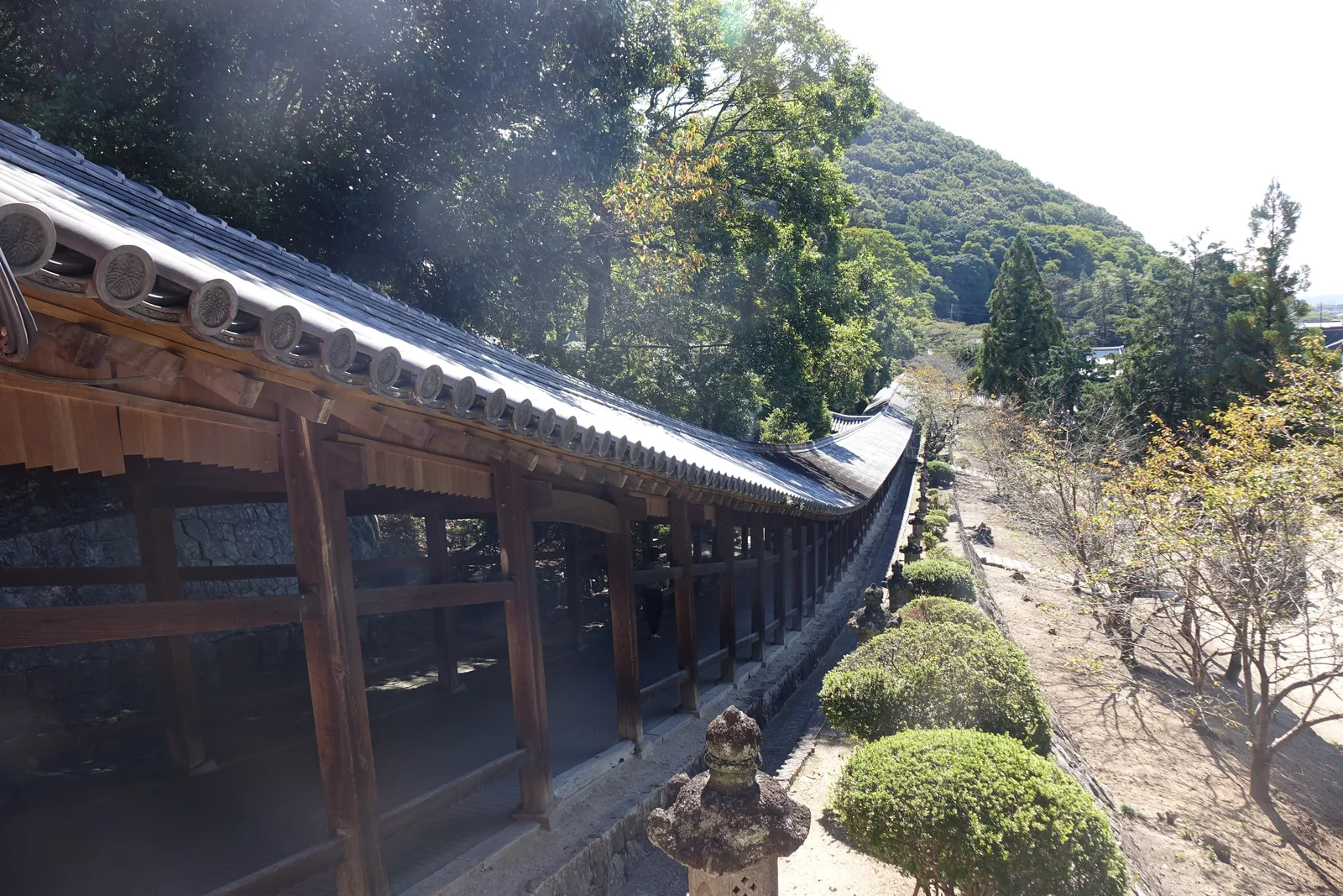從高處看吉備津神社的迴廊：木造的廊子順著山腳的起伏一直線往南延伸，右側是吉備の中山的山體，左側是境內的樹與燈籠