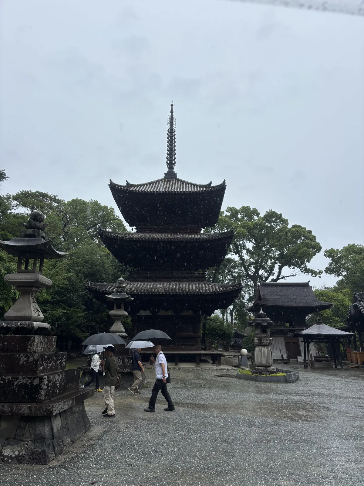 雨中的三重塔。塔前是石燈籠與撐傘走過的參拜者，右側屋簷是另一棟堂宇。這座三重塔是三間四方的和樣建築，明治四十年被指定為重要文化財。