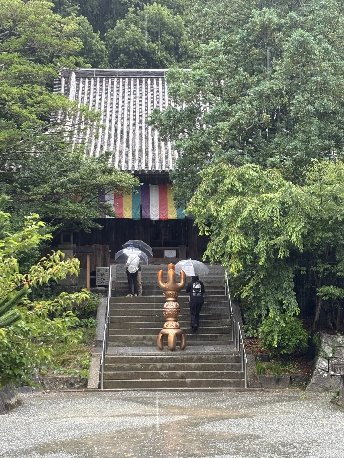 二王門之後往上的石段，正中央立著一具金色的三鈷杵。石段盡頭掛著五色幕，兩個撐傘的人正走上去。