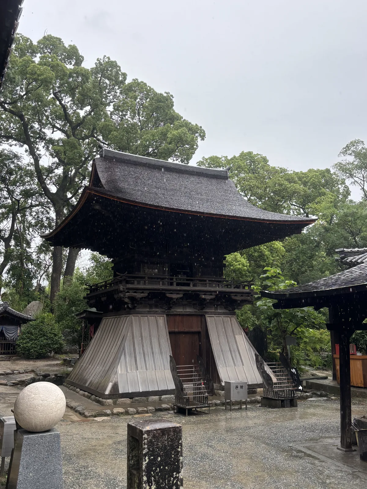 明治四十年指定的鐘樓。下段是袴腰——用木板包成外撇的裙狀，上層有高欄與連子窗，入母屋造本瓦葺。雨把整片瓦打成深色。