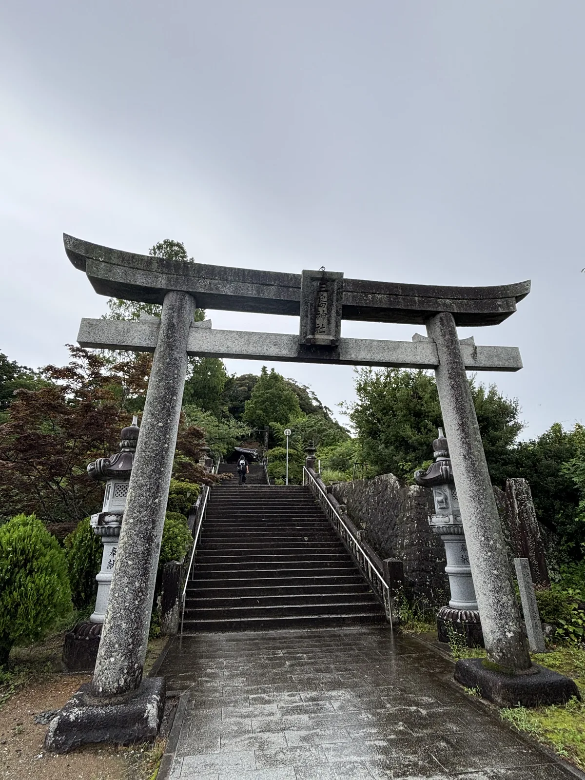 石造明神鳥居與其後直上的石段。額束刻著「三島神社」，左右各一基石燈籠，基座上有奉獻的字樣。石段沒有折彎，兩側被玉垣與樹叢夾住，爬的時候只看得到前方的天空。