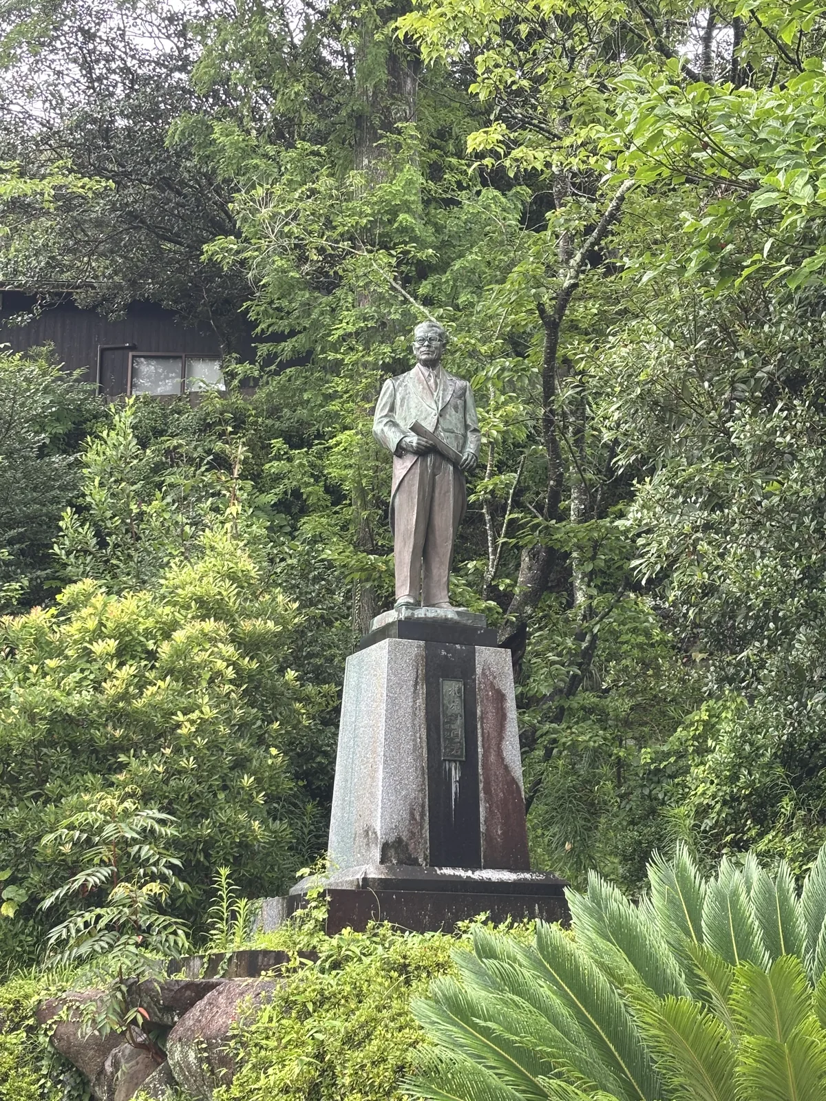 石段下方樹叢裡的西裝銅像，手裡捲著一份文件。台座上的銅牌只刻五個字：「梶原計國君」，沒有生卒，也沒有職銜。