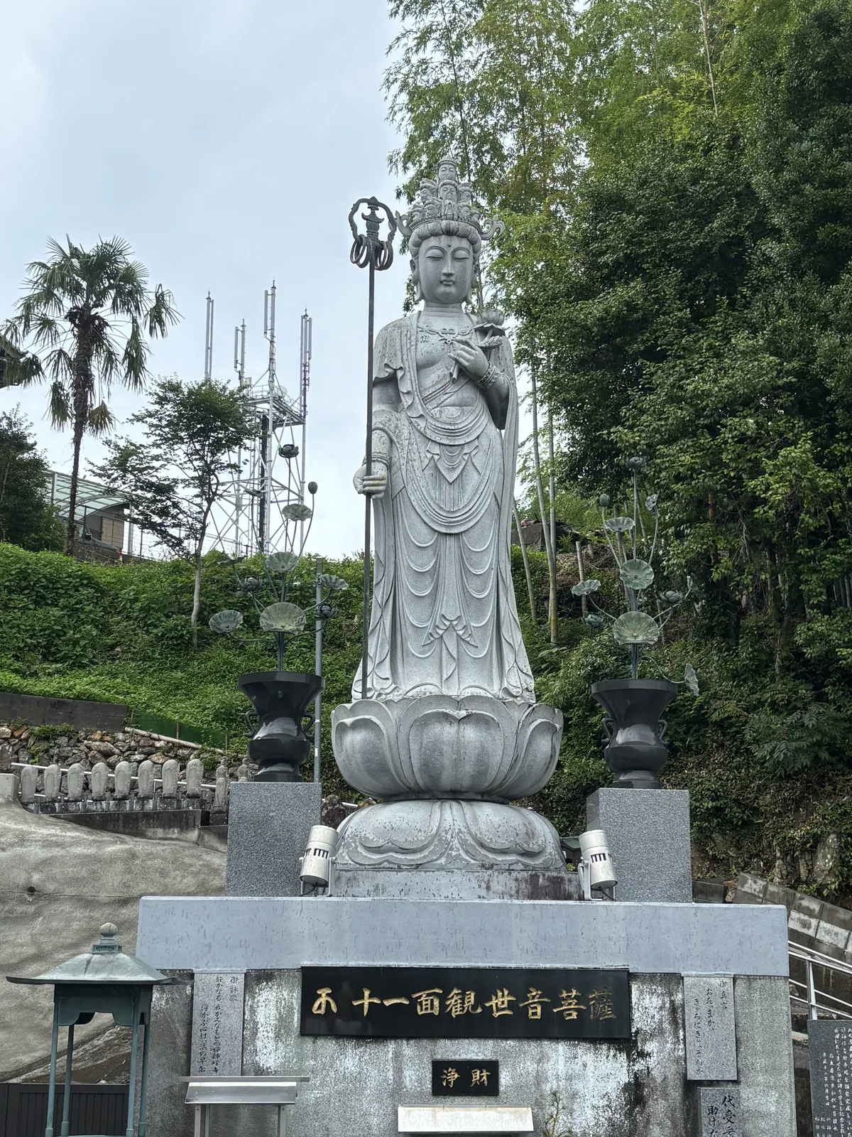站在仰蓮台座上的石造十一面觀世音菩薩立像。黑底金字的台座刻著「西國十一面觀世音菩薩」，下方另有一塊「浄財」。背後是棕櫚與通信鐵塔。