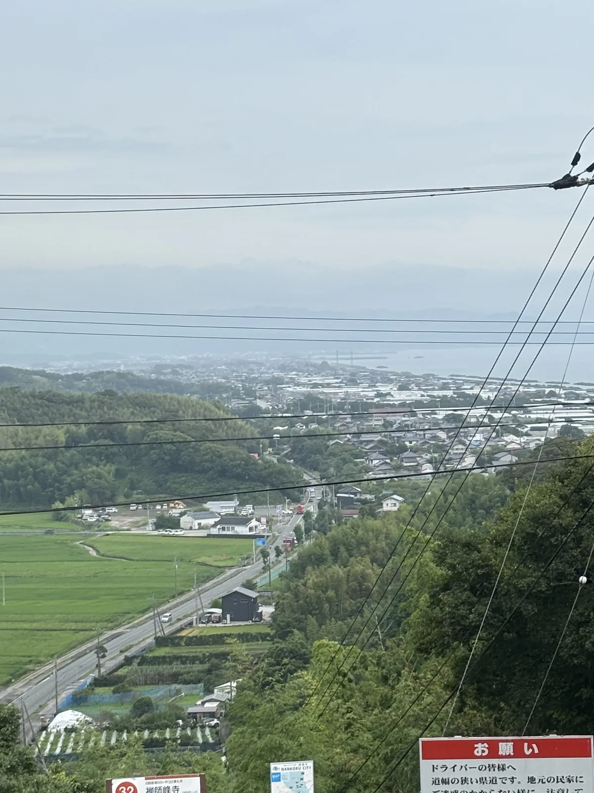 山腳下的水田、溫室與縣道。畫面下緣有一塊紅色的「お願い」（o-negai，請託）告示牌。