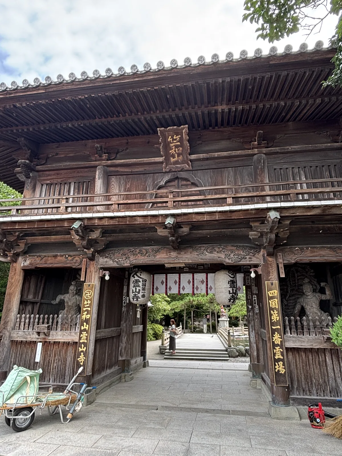 仁王門。木頭已經全黑，正面扁額寫「竺和山」，左右兩塊木牌是「竺和山一乗院霊山寺」與「四国第一番霊場」，兩側的間內站著仁王像，簷下掛著提燈。門前停著一台獨輪車與一綑稻草——那天寺方在整理。
