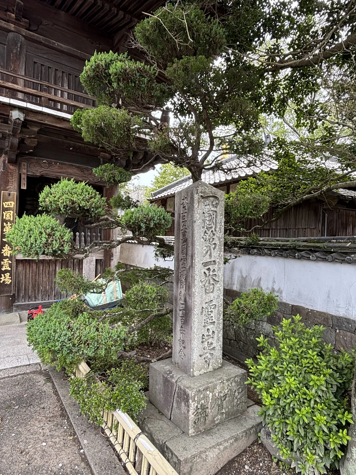 門旁的石標，正面刻「四國第一番靈山寺」，側面刻「四國靈場開創一千二百年記念」。旁邊是修剪成層的圓柏，背後是白牆瓦頂的築地塀。