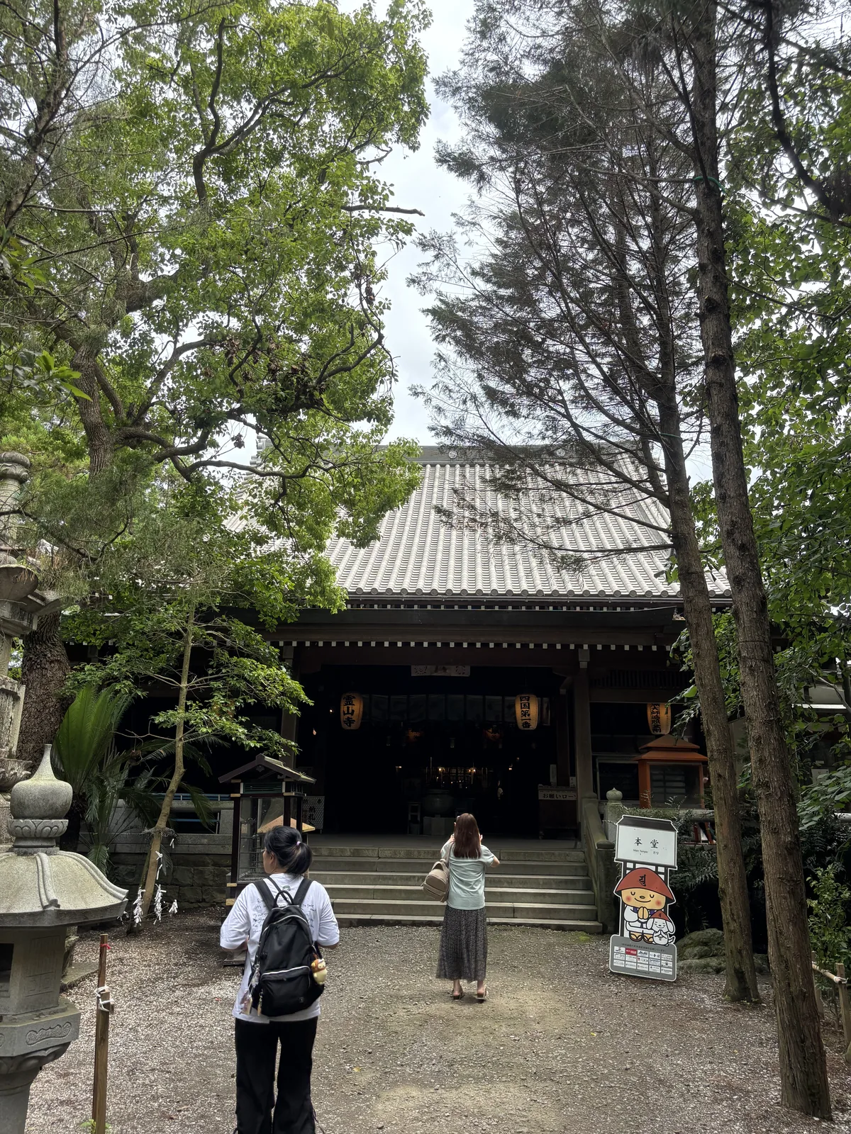 碎石庭、石燈籠、石階；本堂正面全開，堂內深處一片暗。右側立著一具卡通造型的立牌。
