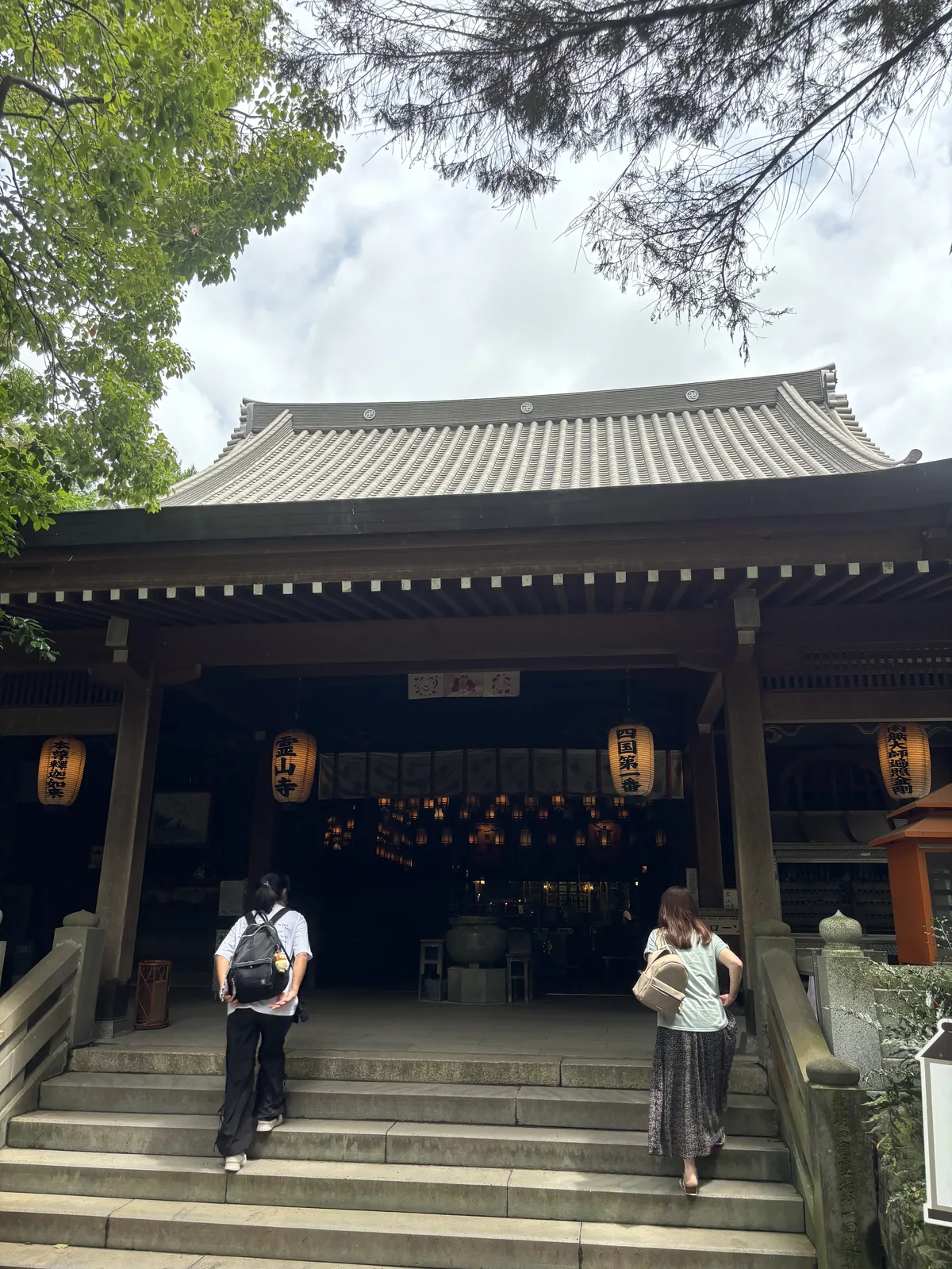 石階上兩排提燈，可讀出「霊山寺」「四国第一」等字；堂內的吊燈籠已經看得見。兩個人正走上階。