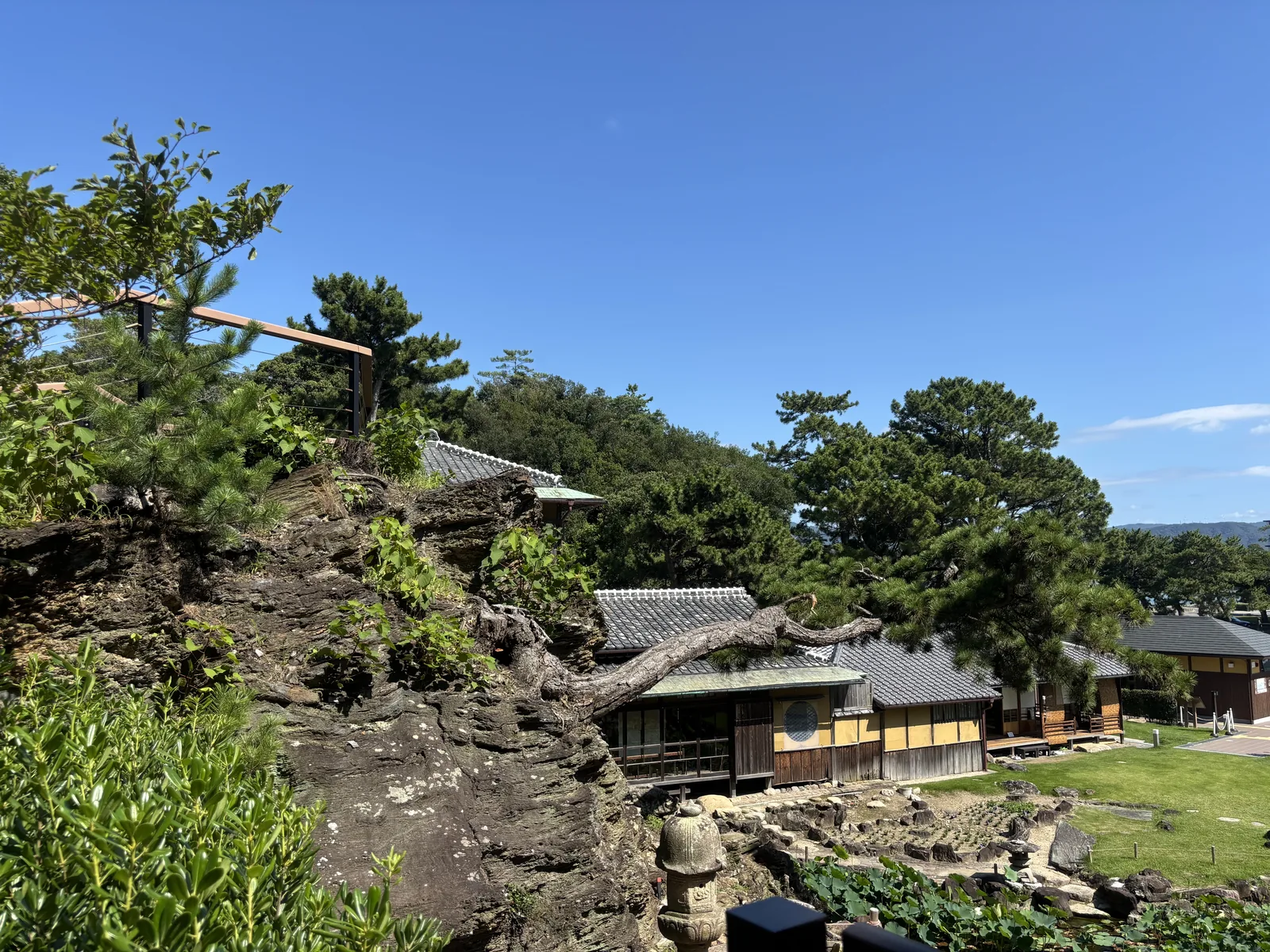 從神社背後的岩山下段往下看：前景是裸露的岩與一具石燈籠，下方是瓦屋頂與草地，遠處是山。六個島剩下的三塊岩，其中一塊就在腳下。