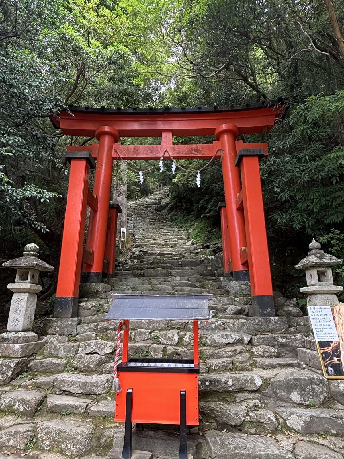 朱色鳥居底下，石段直接開始。兩座石燈籠分立左右，鳥居後方那一片斜上去的灰色就是五百三十八階的第一段。沒有踏面統一的高度，也沒有扶手。