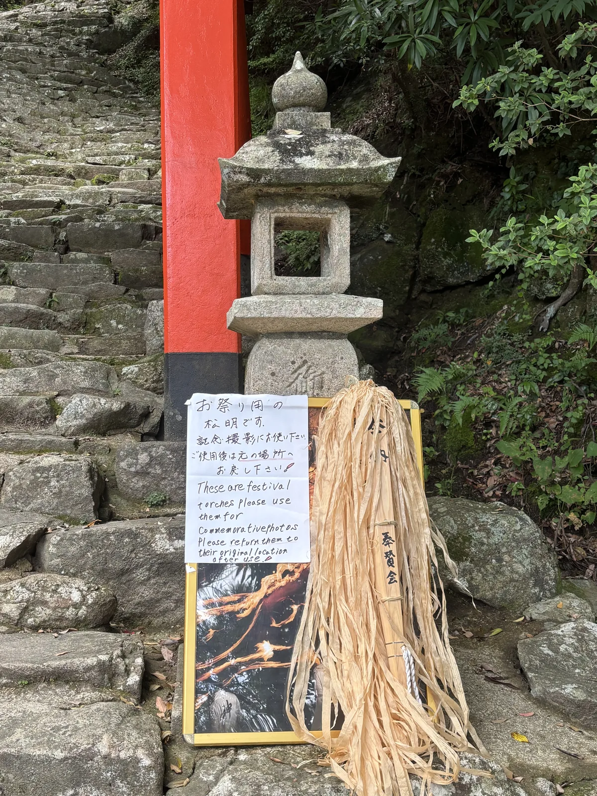 靠著石燈籠立著一疊真的稻稈松明，綁著「奉賛会」的木牌。旁邊手寫的紙日英並排：「お祭り用の松明です。記念撮影にお使い下さい。ご使用後は元の場所へお戻し下さい。」
