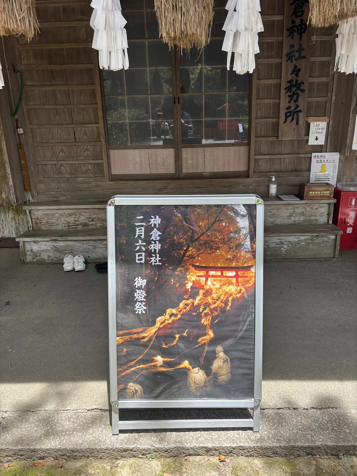 掛著注連繩與紙垂的木造小屋。門口一塊壓克力立牌，照片是火從石段上流下來的樣子，標題「神倉神社　二月六日　御燈祭」。御神札與御朱印平時在熊野速玉大社受理。
