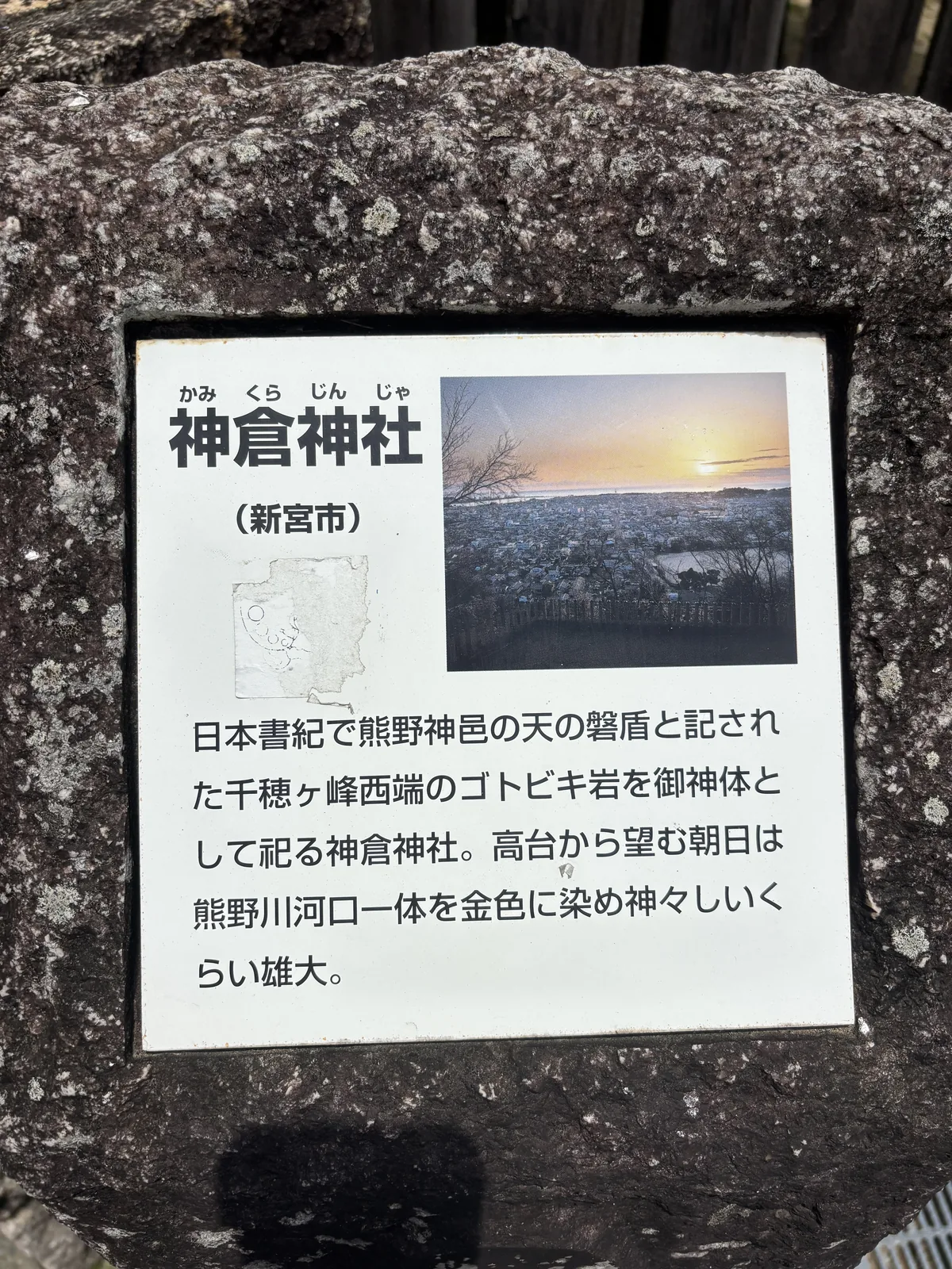 嵌在一塊粗糙巨石上的白色說明板：「日本書紀で熊野神邑の天の磐盾と記された千穂ヶ峰西端のゴトビキ岩を御神体として祀る神倉神社。高台から望む朝日は熊野川河口一体を金色に染め神々しいくらい雄大。」右上角附的照片就是那個日出。