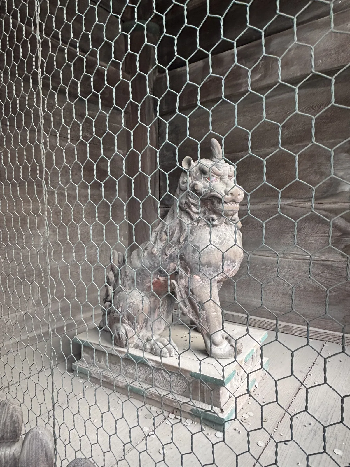 六角鐵網後面的石造狛犬（成對的守護石獸），紅色顏料還剩一點，蹲在綠漆的台上。要看清楚得把臉貼到網上。