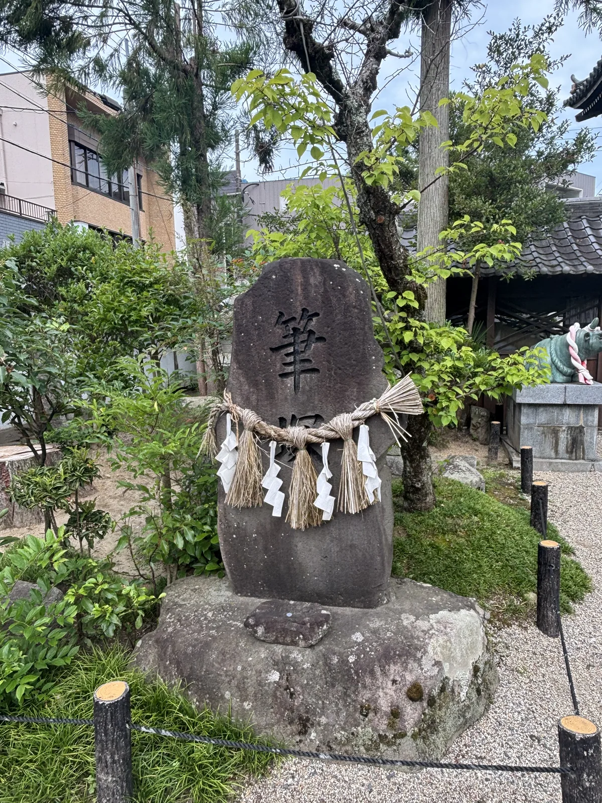 自然石上刻大字「筆塚」——把用舊的筆埋起來供養的塚，纏著注連繩、四束稻藁與紙垂，座在一塊粗石上。右後方屋簷下是青銅的神牛，繫紅白繩，架在花崗岩台座上。學問與農耕，這座神社的兩層來歷各有一件實物。