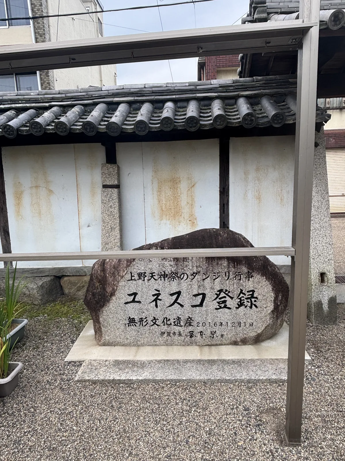 自然石刻「上野天神祭のダンジリ行事／ユネスコ登録／無形文化遺産　2016年12月1日」——上野天神祭的花車行事，聯合國教科文組織登錄，非物質文化遺產。落款是伊賀市長。碑立在土塀前，上頭罩著鋼構的雨遮。