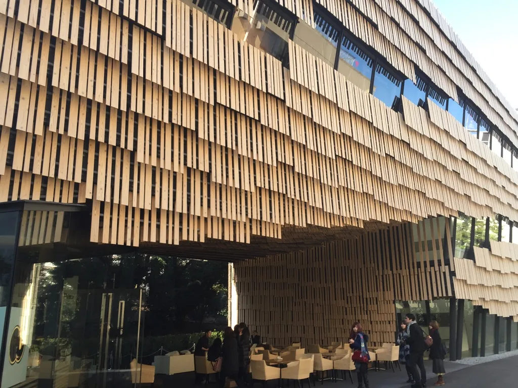 大和泛在學術研究館東側外觀，三層高的立面覆滿長短不一的杉木板，木板在一樓處向內捲成懸吊的天花，下方是咖啡座與穿越建築的通道