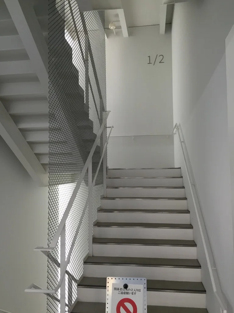 研究館內部的樓梯，白色的鋼製樓梯沿著白牆上升，一側以穿孔金屬板圍合，牆上標著樓層的數字，梯口立著禁止進入的標示牌