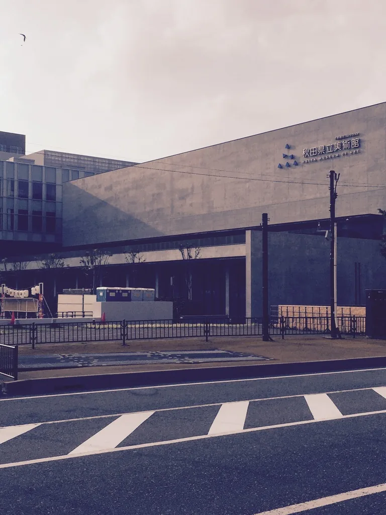 自道路對側望美術館，清水混凝土的量體頂部掛著館的標誌，下層後退，前方是護欄與斑馬線