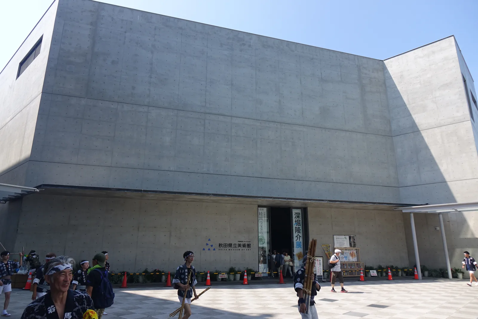 竿燈祭當天的美術館正面，上層的清水混凝土牆面向兩側轉折，下層後退設有入口，廣場上聚集著身穿法被的人群