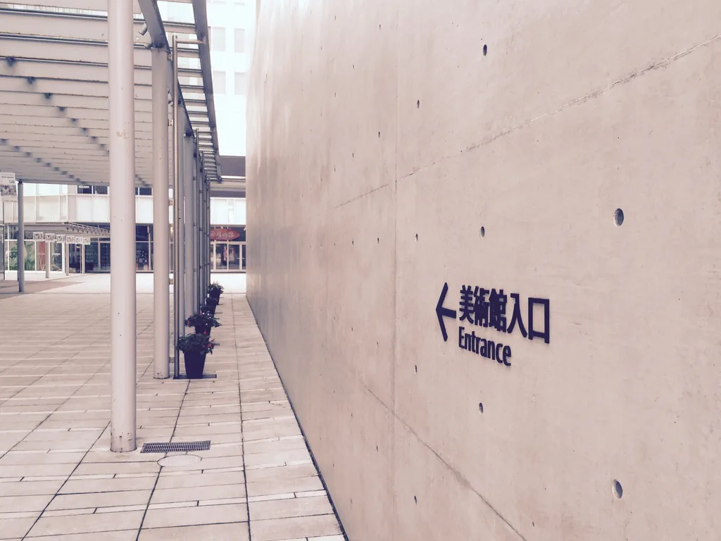 清水混凝土牆上以黑色字標示美術館入口與英文 Entrance，左側為細柱支撐的雨庇與鋪石步道