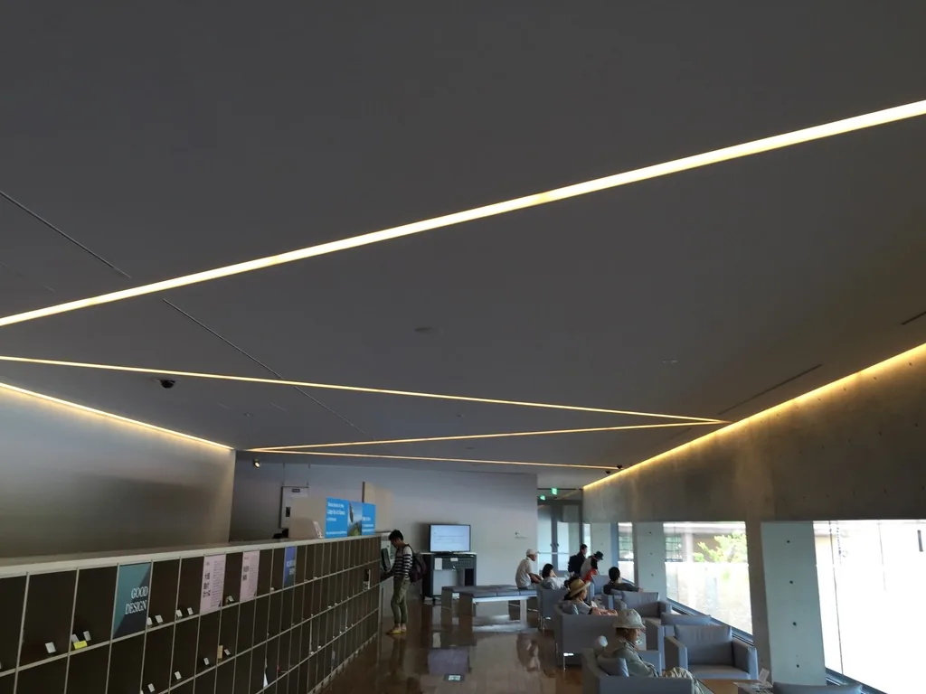 museum lounge 的縱深，左側沿牆是低矮的書架，右側是面向水庭的玻璃，深灰色的天花以斜向的線形燈具分割，座位上有訪客