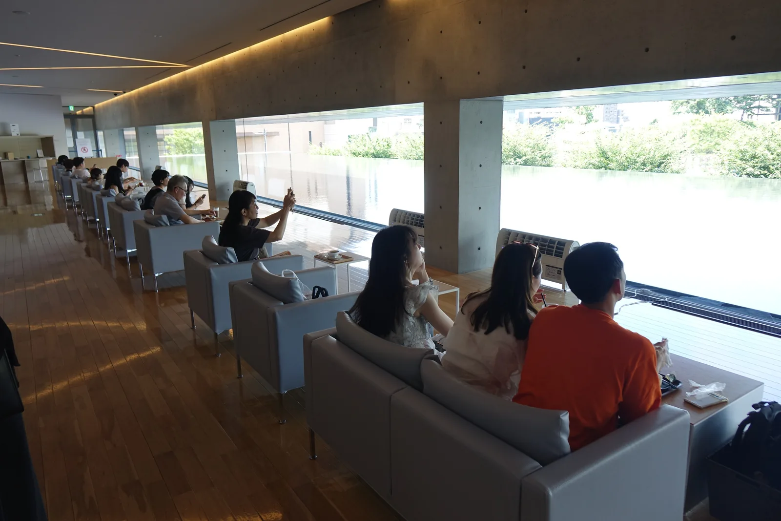lounge 沿玻璃排列的沙發座位，訪客面向窗外，窗外是水庭與對岸的綠意，天花的線形燈具沿空間方向延伸