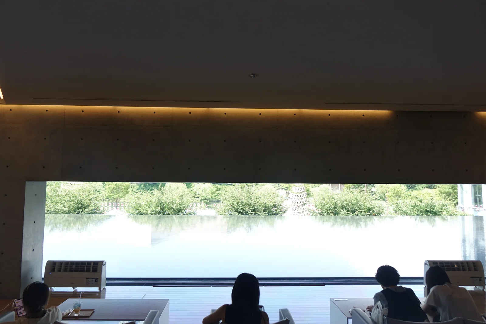 lounge 的低開口，上緣是一道深色的水平梁與間接照明，下方是水庭與對岸的綠樹，前景為訪客的剪影與桌椅