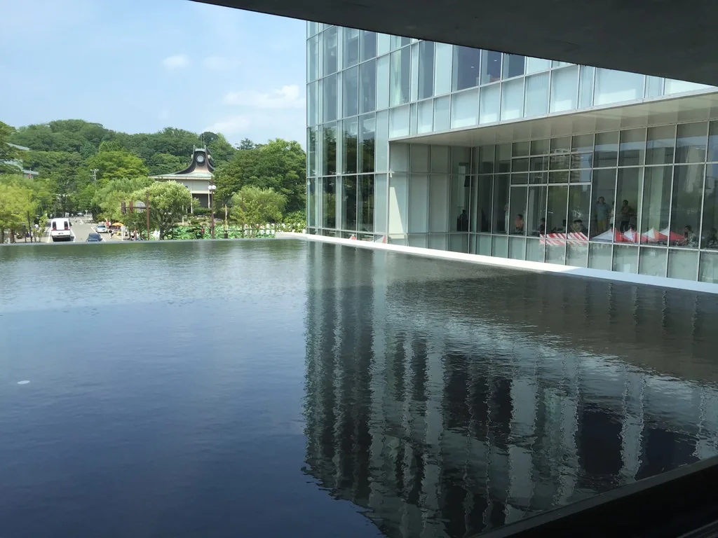水庭的水面向遠處延伸，右側是 lounge 的玻璃量體與倒影，遠方是外堀對岸的樹林與一座帶塔的建築