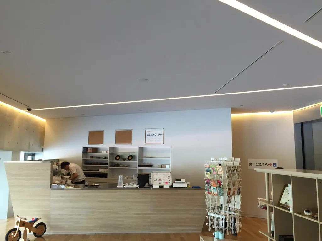 lounge 一角的咖啡吧台，吧台以淺色木紋面材包覆，後方牆上是展示架與點餐處的告示，前方放著明信片架與一輛小型木製腳踏車
