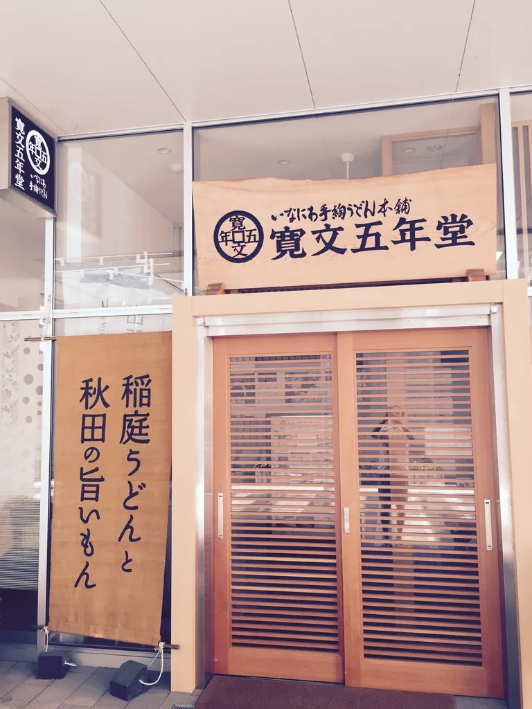 寬文五年堂秋田店的入口，木製格柵拉門上方掛著店名招牌，左側立著寫有稻庭烏龍麵與秋田美味的布幔